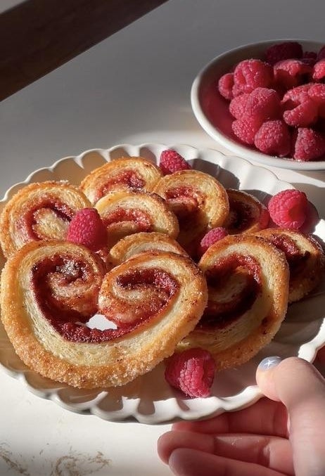 Pastry hearts with raspberry filling connected  a plate, garnished with caller  raspberries. Hand reaching towards the dish