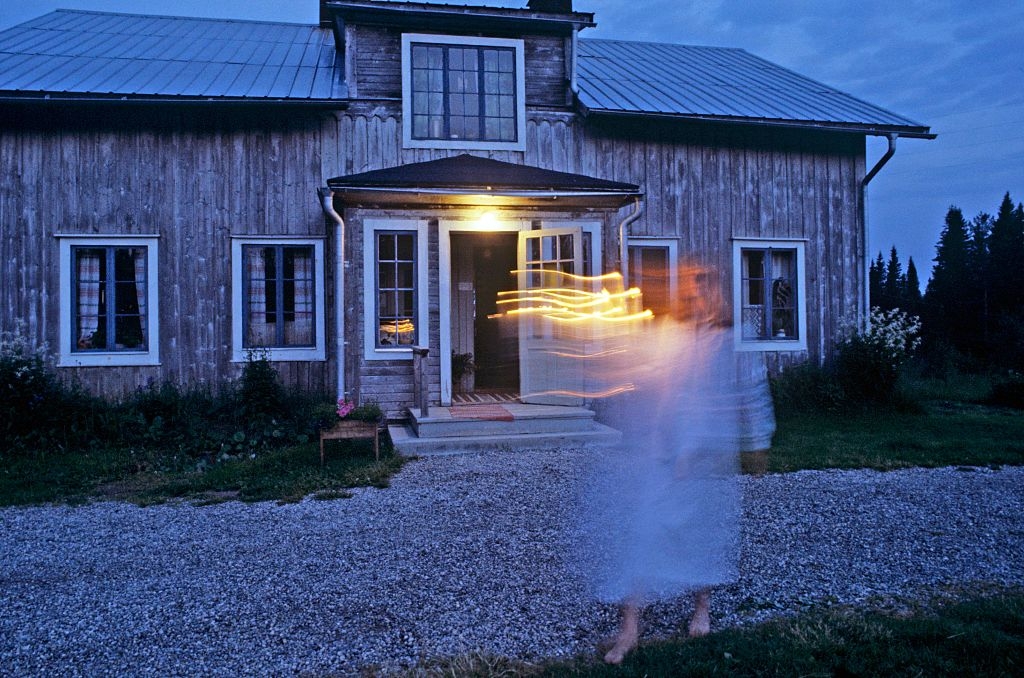 A blurred, ghostly fig successful a achromatic robe stands successful beforehand of a rustic woody location astatine dusk, with a lukewarm airy glowing from the doorway