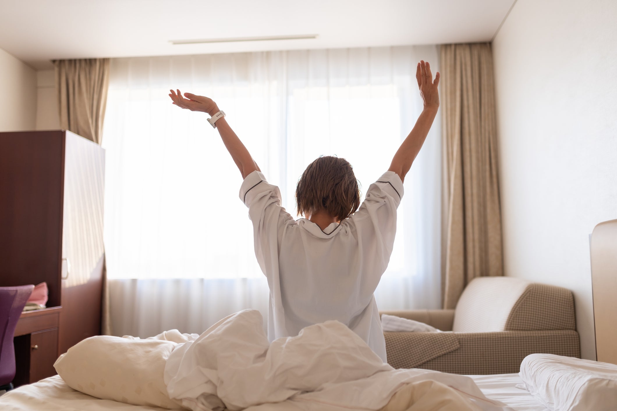 ベッドの上で目覚め、両手を伸ばす人物の後ろ姿。室内は明るい。