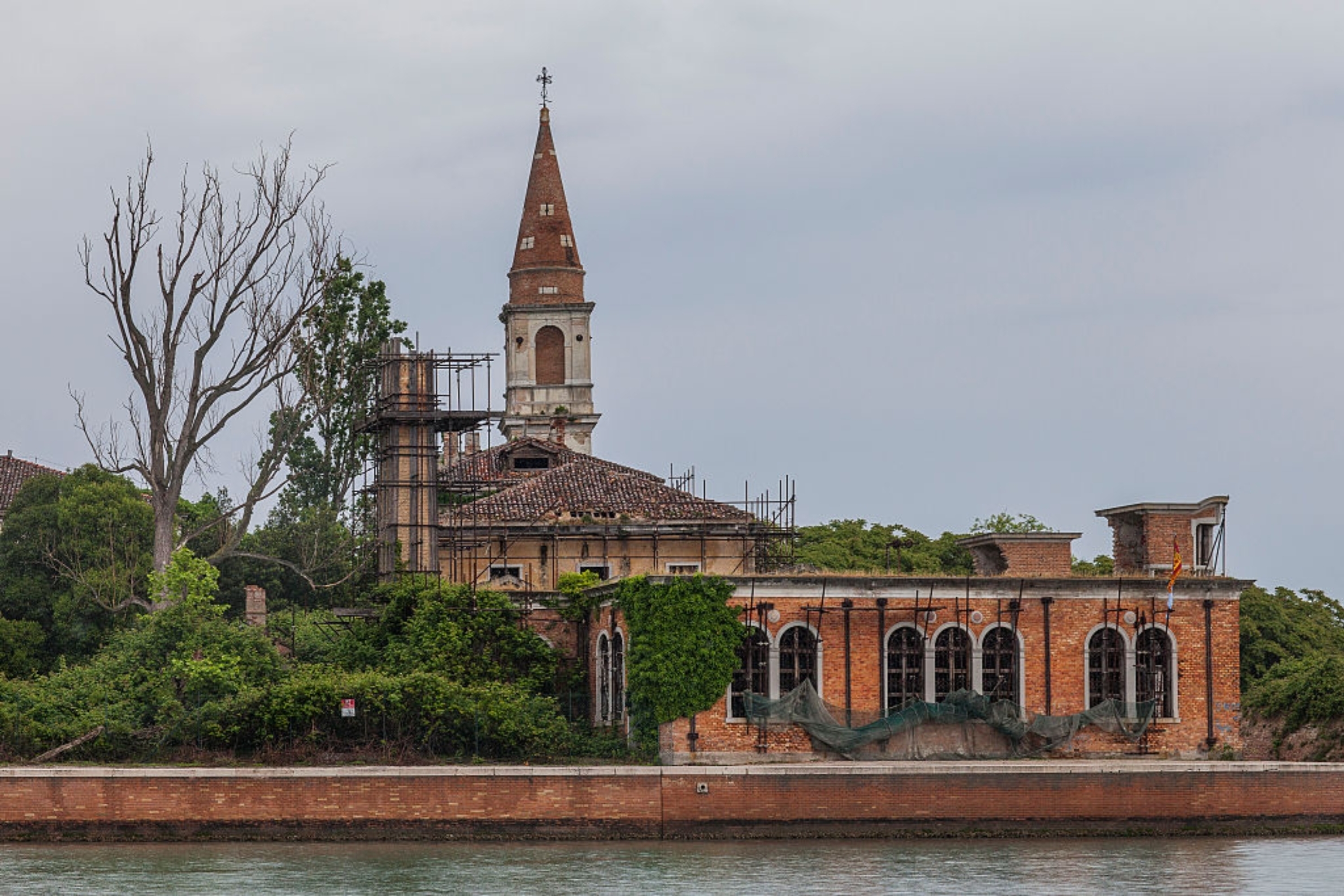 Old, partially renovated gathering with a operation and scaffolding, surrounded by trees, adjacent water