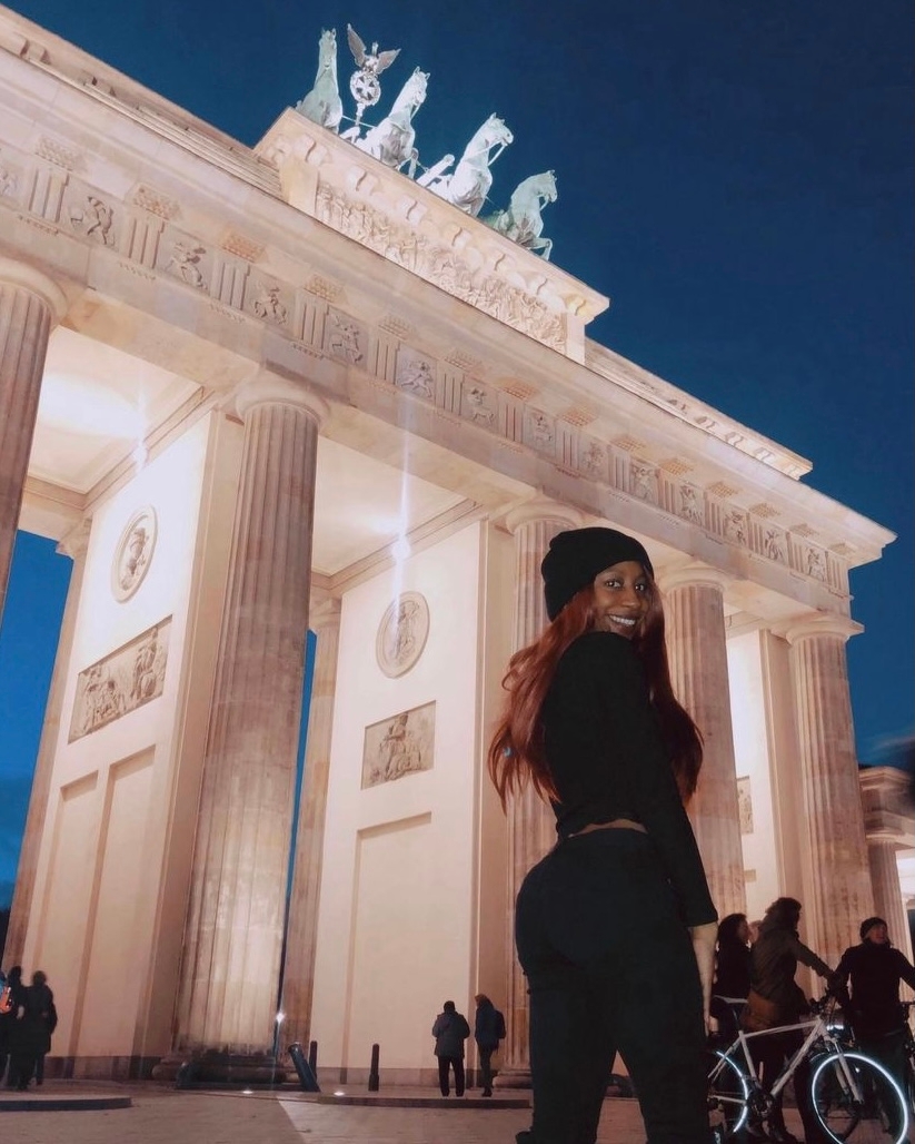 Person stands smiling near Berlin's Brandenburg Gate, wearing a beanie and dark outfit at dusk