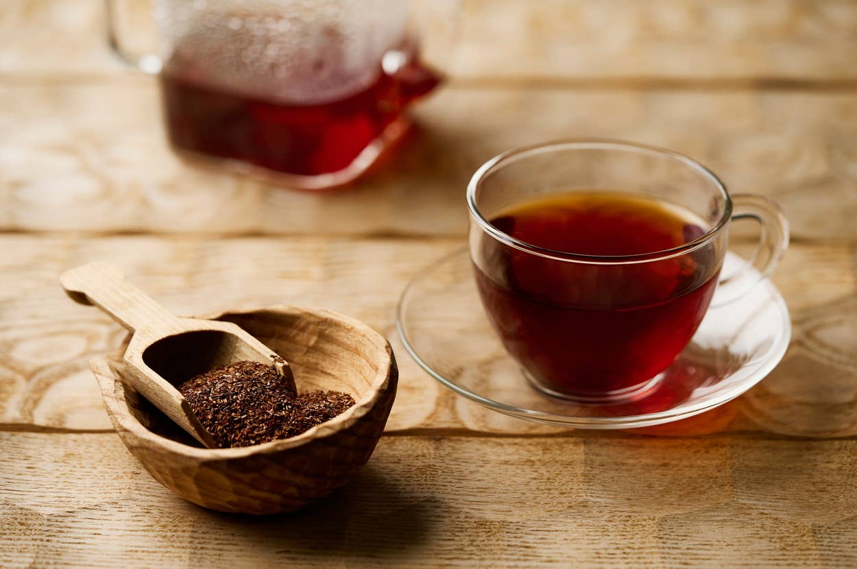 A glass cup of tea sits on a table next to a wooden bowl and scoop filled with loose tea leaves