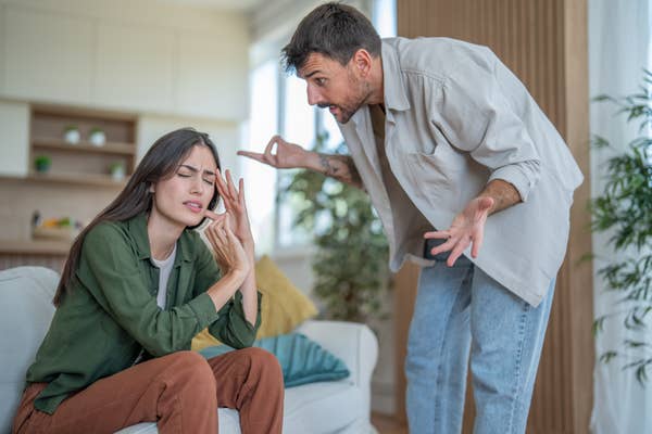 Man angrily gestures at woman sitting on couch, looking distressed with eyes closed and hand on forehead