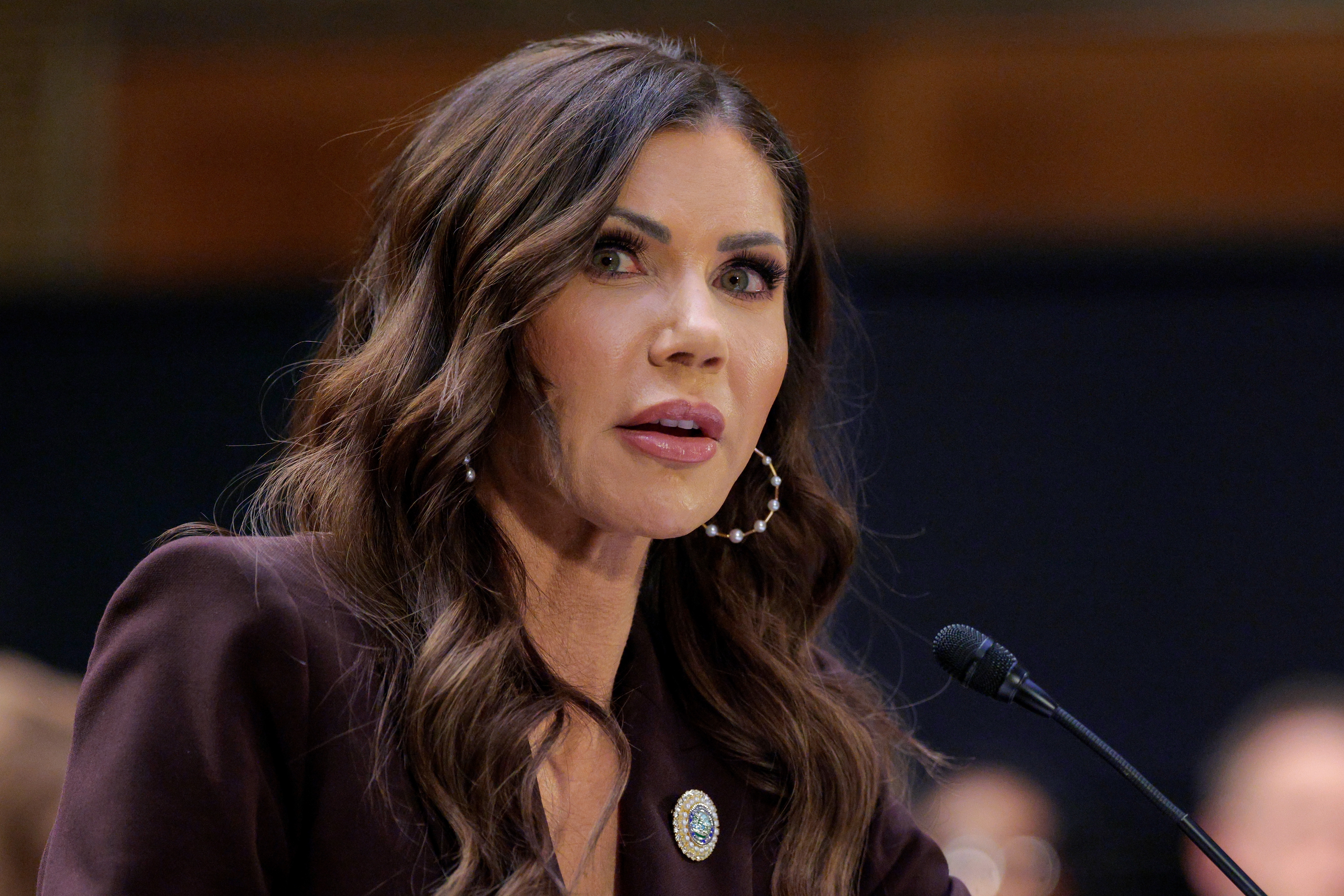 A pistillate   with long, wavy hairsbreadth  speaks astatine  a microphone during a ceremonial  event, wearing a suit   with a circular  pin