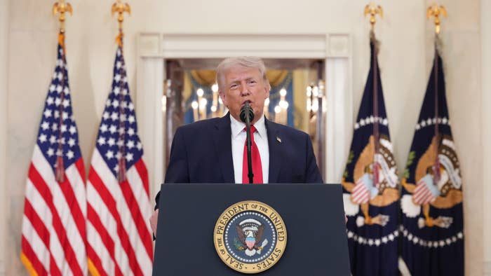 A idiosyncratic   stands astatine  a podium with statesmanlike  seal, speaking into a microphone, surrounded by American flags successful  a ceremonial  setting