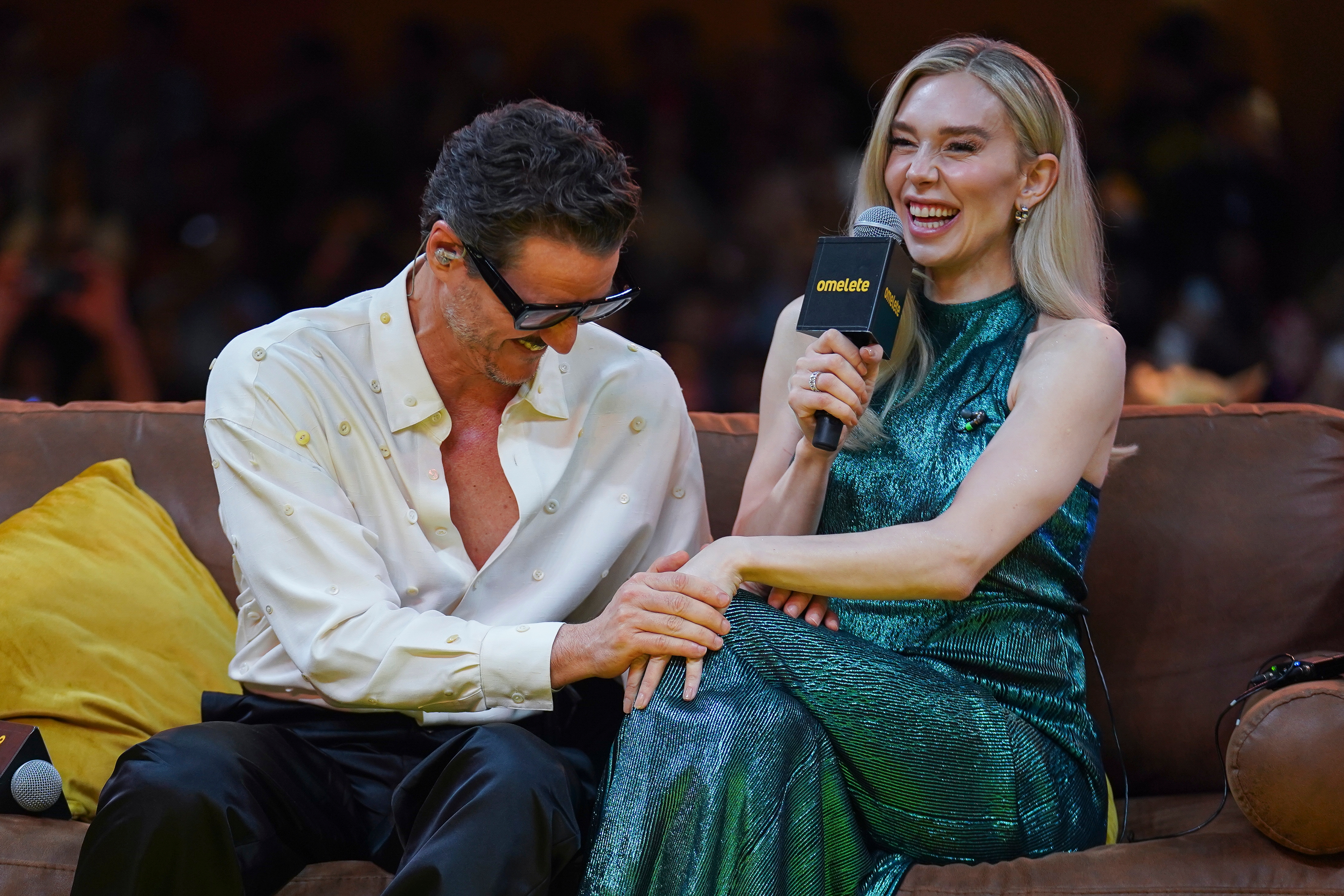 Two celebrities seated connected  a sofa, laughing. One is wearing a shiny, textured gown, the different   successful  a casual open-collared garment  and trousers