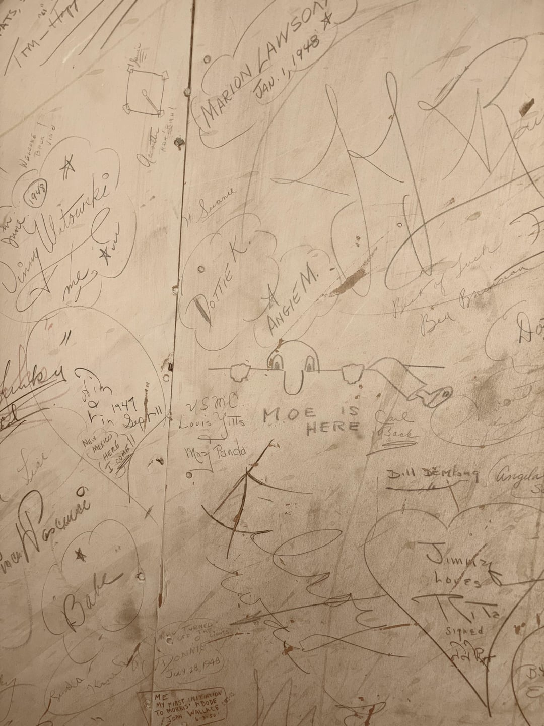 Graffiti-covered partition  with assorted  signatures and doodles, including "Moe is here" successful  the center. Various names and dates are visible