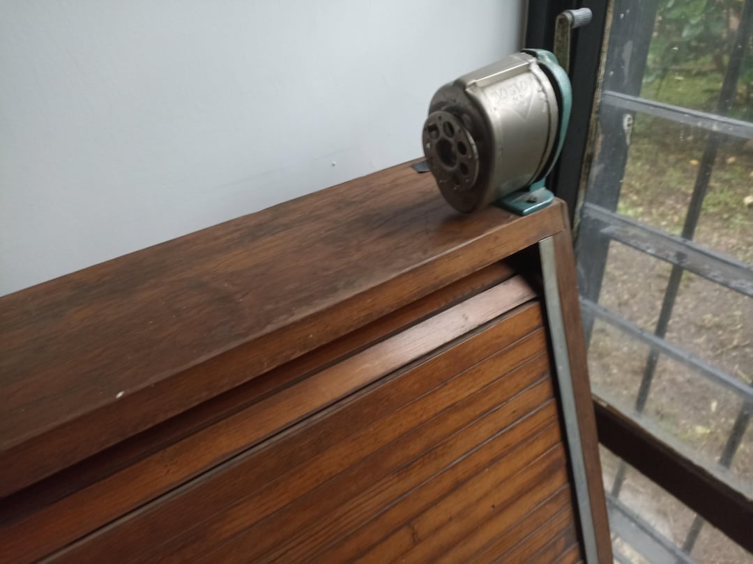 A vintage pencil sharpener mounted connected  a woody  schoolroom  desk, adjacent   a window