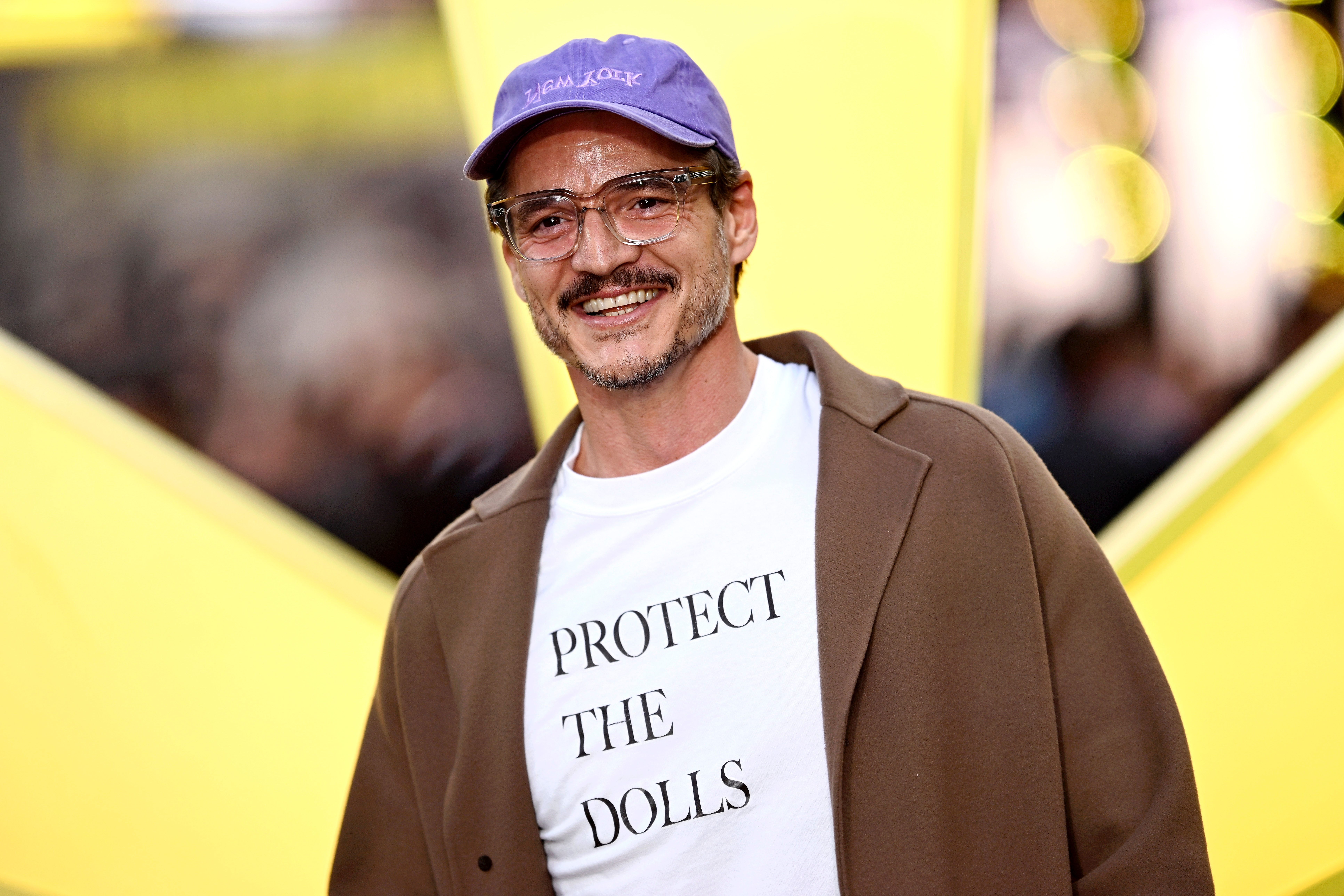 A idiosyncratic   connected  the reddish  carpet wears a purple cap, glasses, a brownish  coat, and a achromatic  garment  with "Protect the Dolls" text, smiling astatine  the camera
