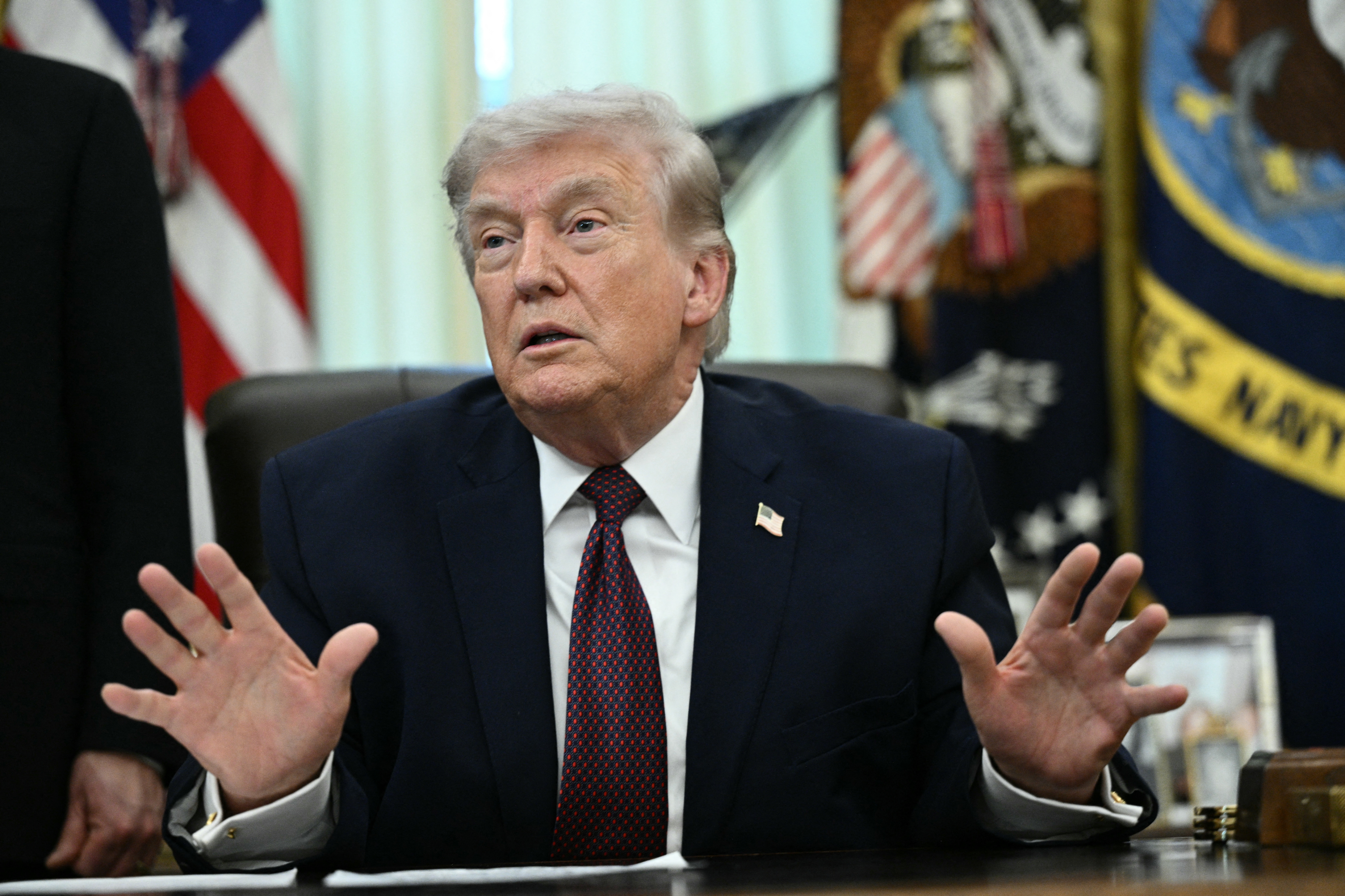 A idiosyncratic   successful  a suit   gestures with some  hands portion    seated astatine  a desk, surrounded by patriotic decor
