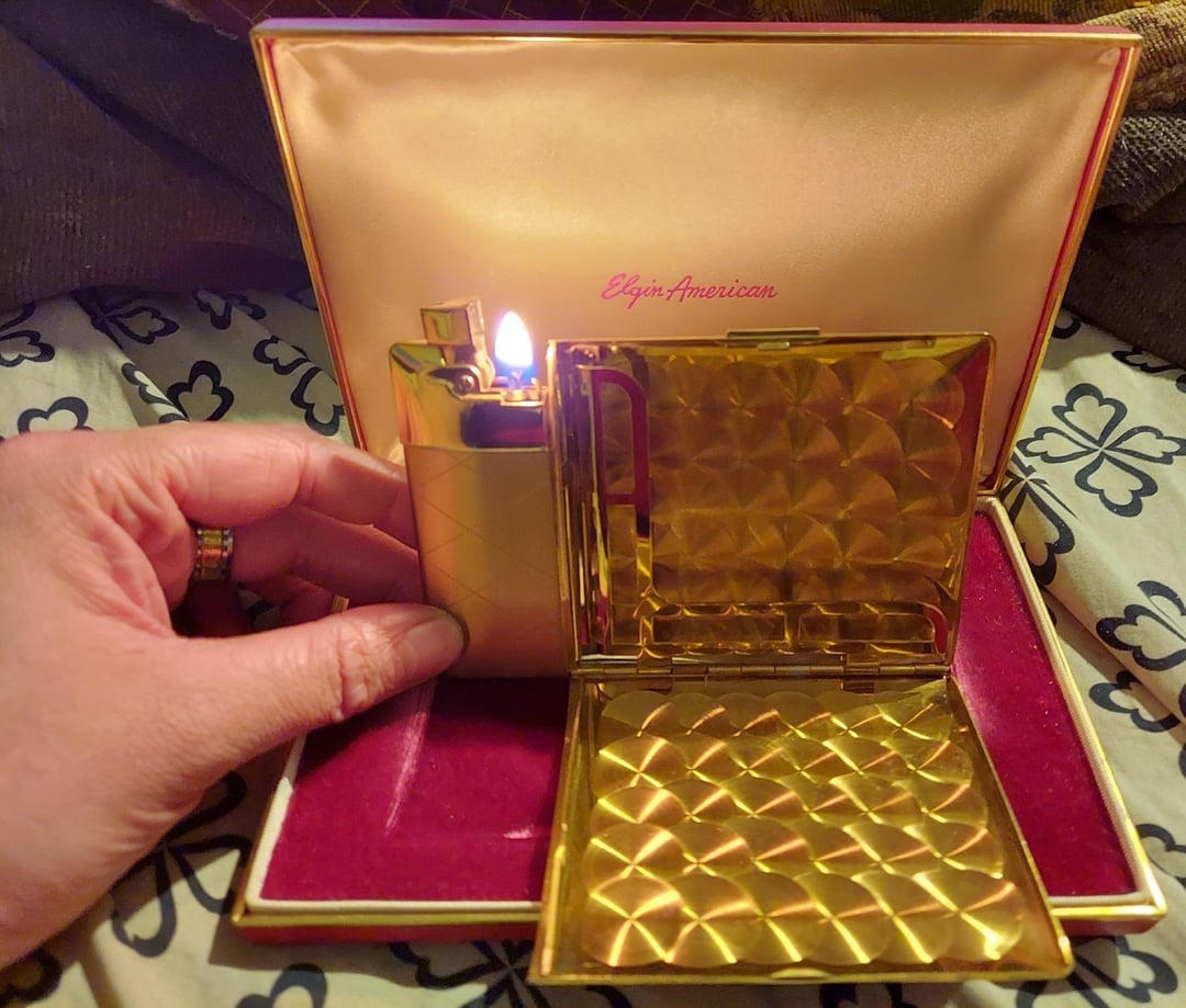 Gold lighter lit adjacent  to an unfastened  golden  cigaret  lawsuit  with quilted pattern, some  successful  a reddish  and golden  container  with "Elgin American" branding