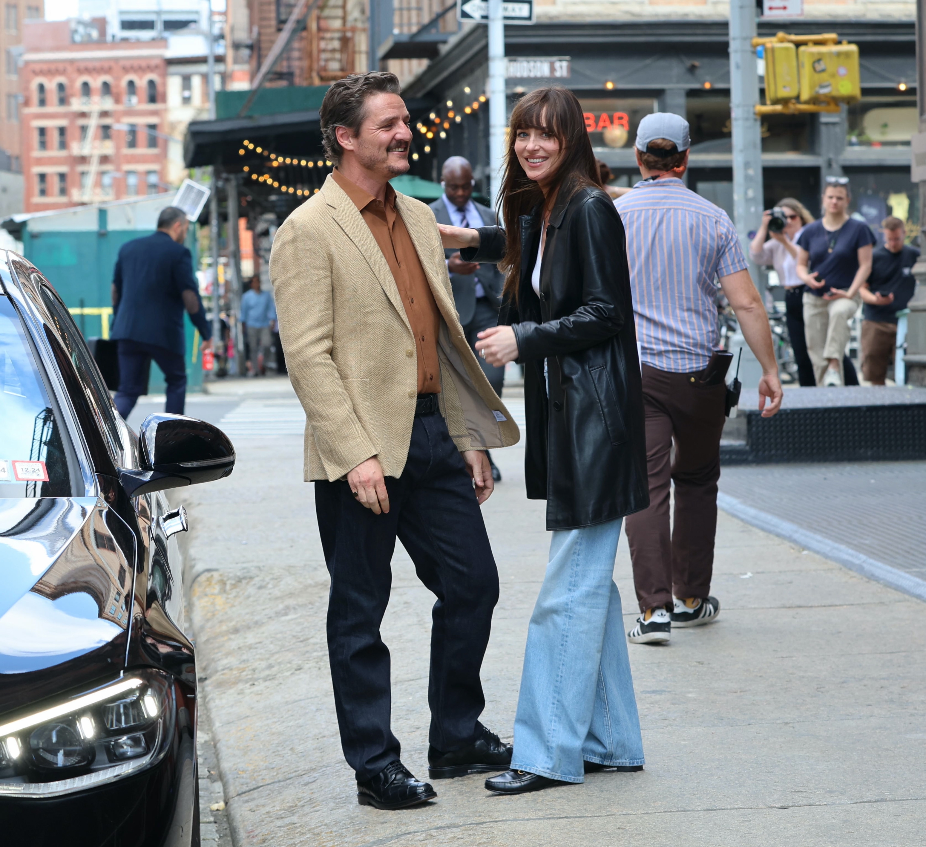 Two radical   smiling and conversing connected  a metropolis  sidewalk; the pistillate   wears a leather overgarment   and wide   jeans, the antheral   is successful  a beige blazer and formal  pants