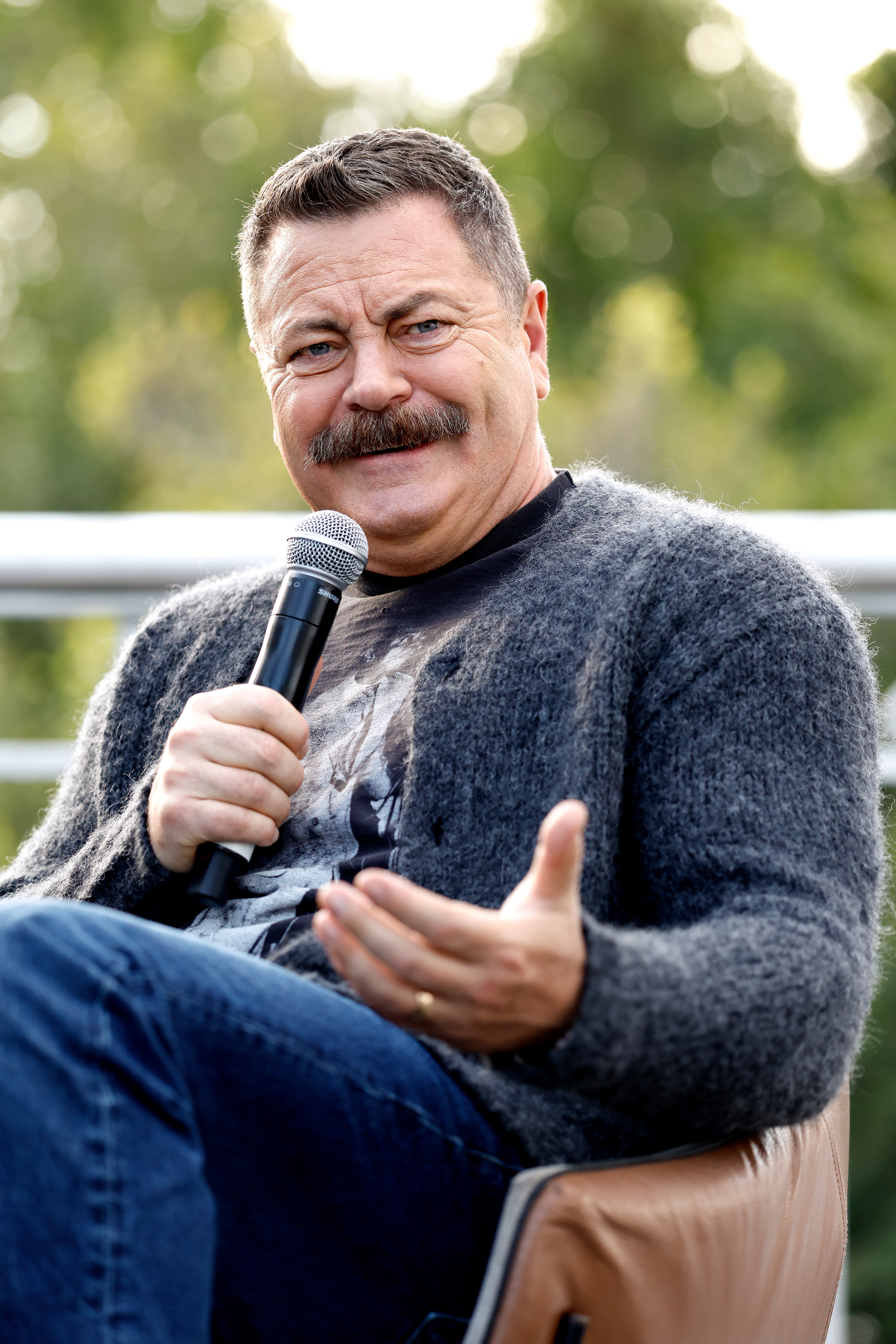 Man holding a microphone, wearing a casual sweater and jeans, gesturing portion    seated outdoors