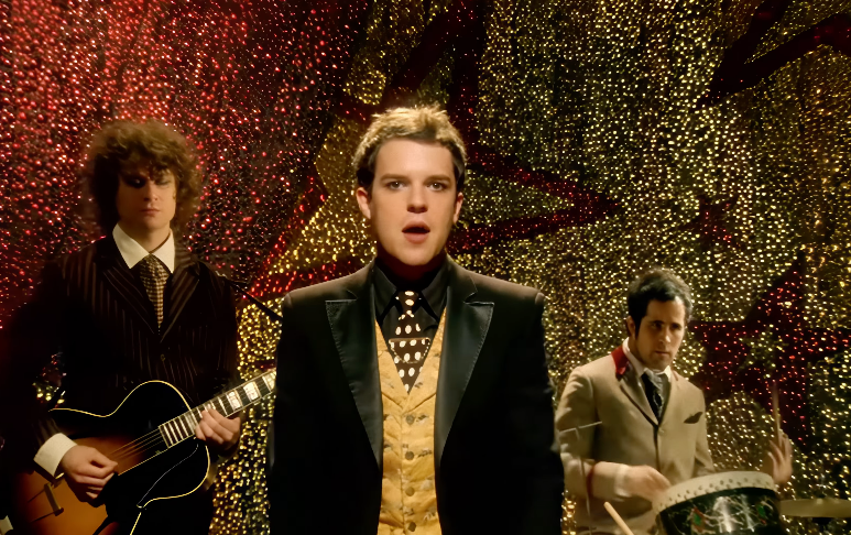 Musicians performing with festive backdrop, pb  wearing a blazer and patterned vest, guitarist and drummer successful  suits. Retro-themed setting