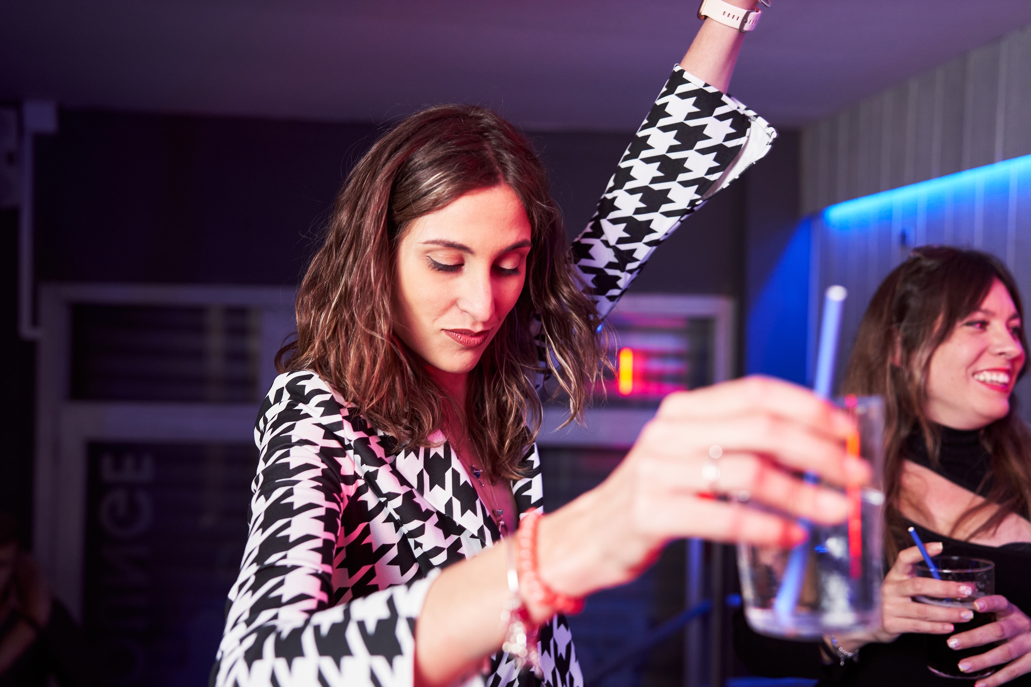 Woman successful a patterned blazer holding a drink, dancing with different idiosyncratic astatine a societal event