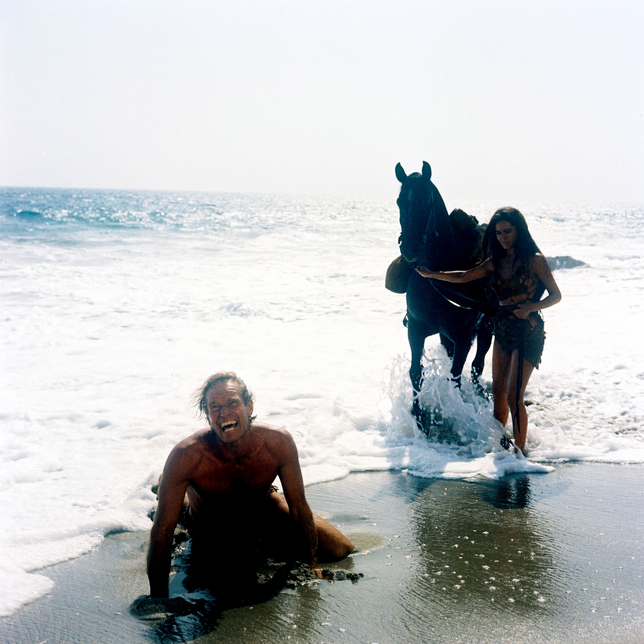 A antheral   kneels connected  a beach, laughing. A pistillate   successful  rugged attire stands beside a equine  successful  the surf. The country   suggests a cinematic moment