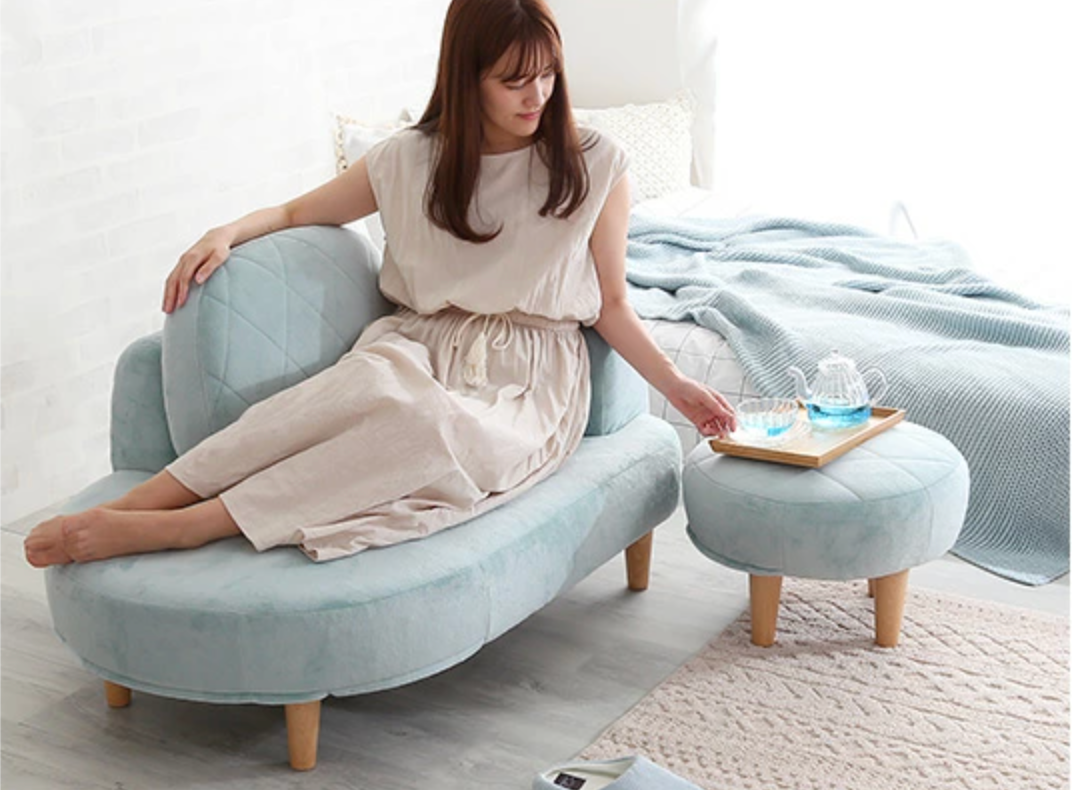 女性がリビングルームでChammy「大人かわいい コンパクトカウチソファ オットマン付き ソファー」に座り、トレイに置かれた飲み物を手で取ろうとしているシーン。