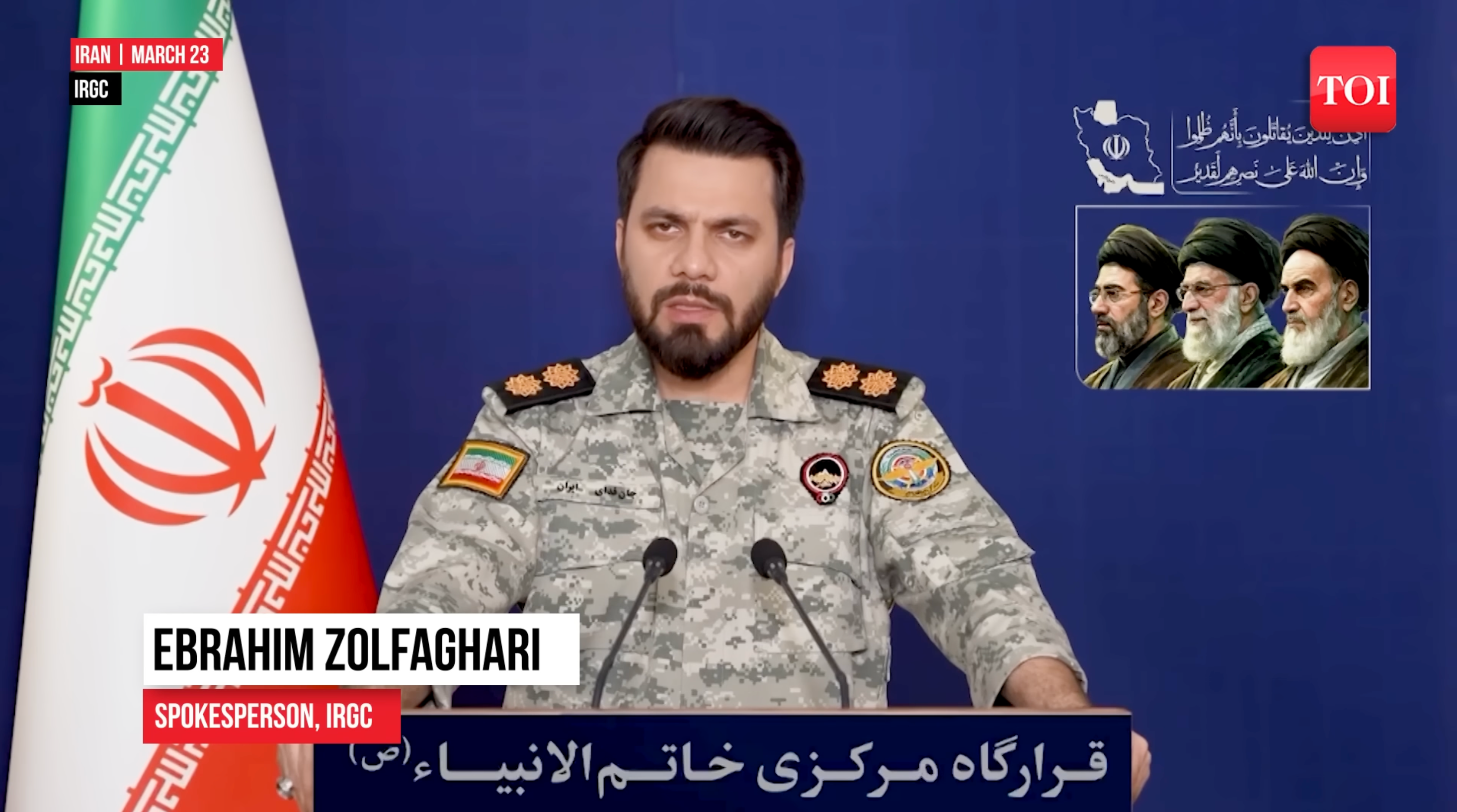 En militær talsperson i uniform henvender seg til media ved siden av et iransk flagg og portretter av ledere