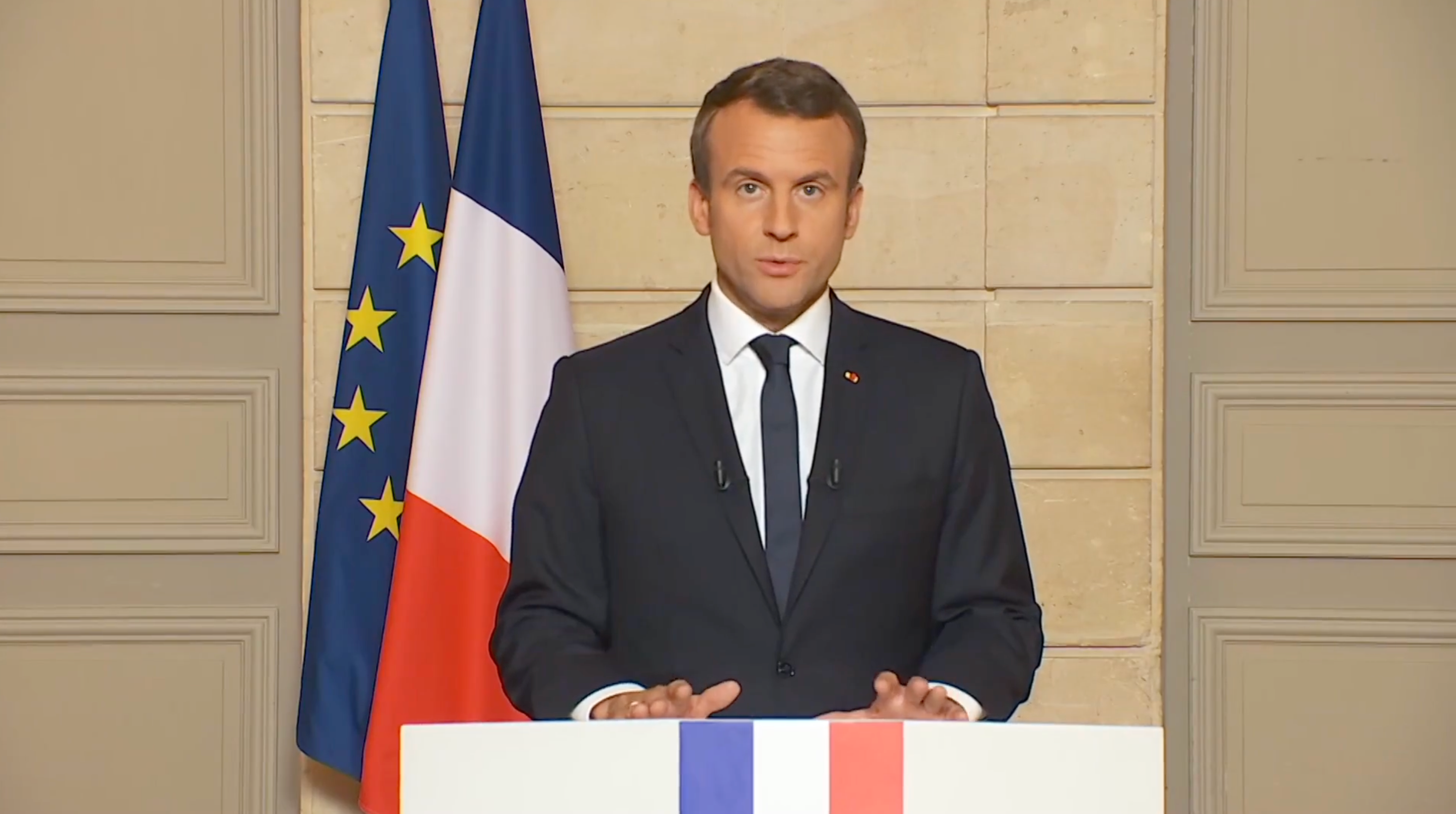 Person som holder en tale på et podium foran franske og EU-flagg, iført en formell dress og slips