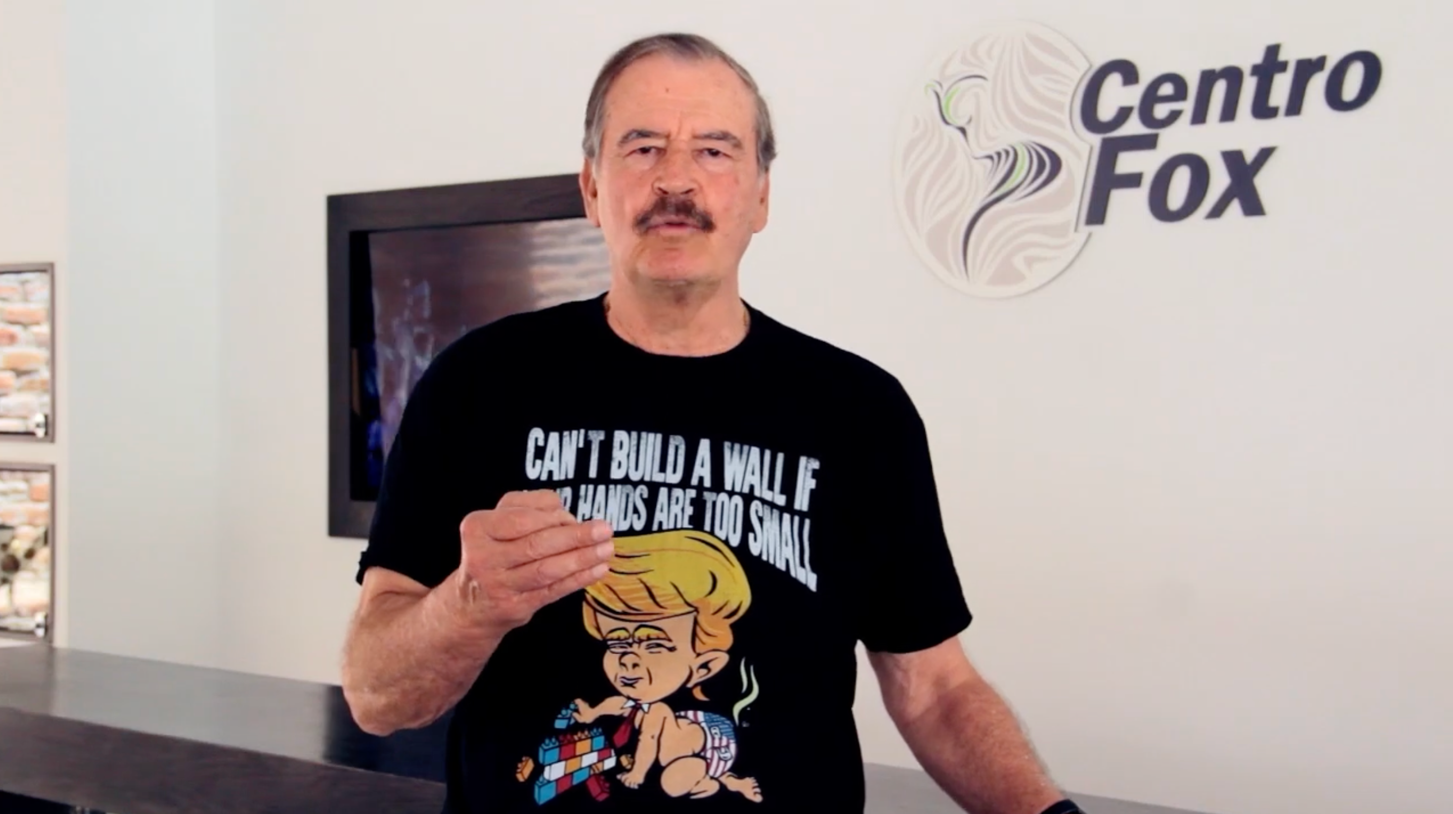 Mann iført en T-skjorte med en tegneserie av en politiker som holder blokker, stående foran en 