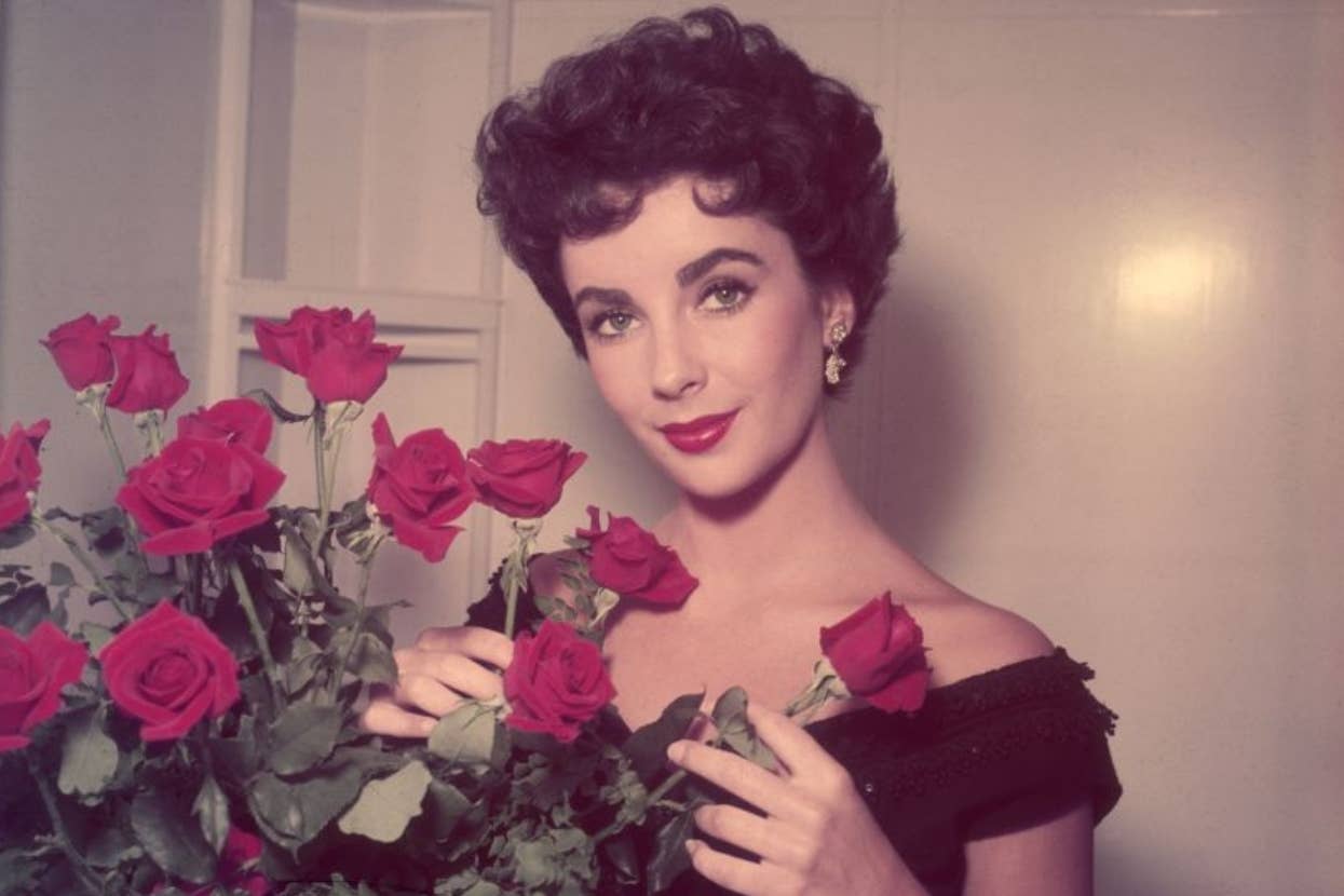 Elizabeth Taylor holding a bouquet of roses, with short, wavy hair, wearing an off-the-shoulder dress and earrings