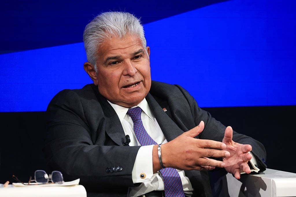 Person i dress og slips snakker på et arrangement, gestikulerer med hendene, sitter på en scene med blått bakteppe