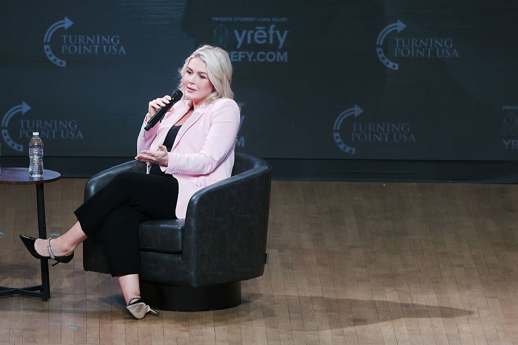 Person som sitter på scenen, snakker i en mikrofon, iført en lett jakke og svarte bukser på et Turning Point USA-arrangement