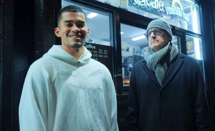 Two radical   lasting  extracurricular  a cafe. One wears a hoodie and smiles; the different   wears a coat, scarf, and hat, looking serious