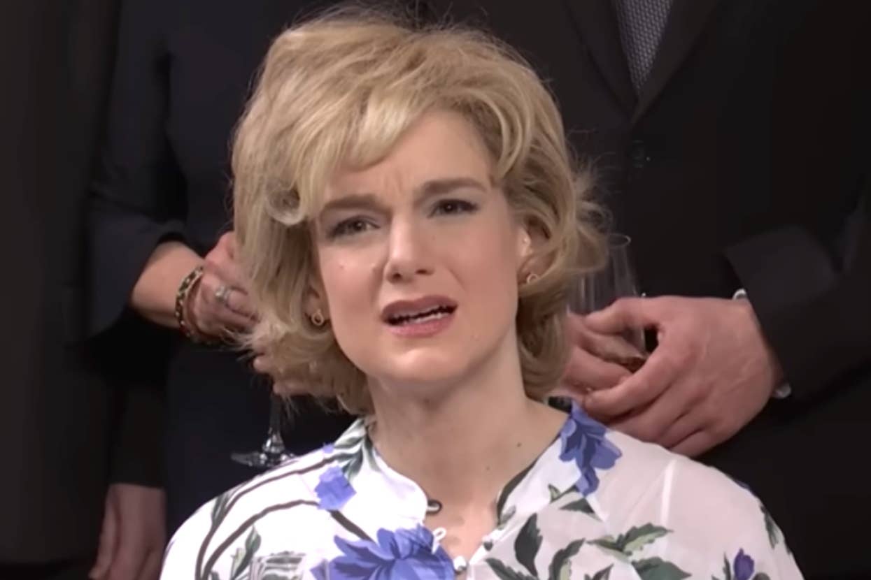 Person with short, styled hair wearing a floral-patterned top, looking forward. Background shows partial figures holding glasses