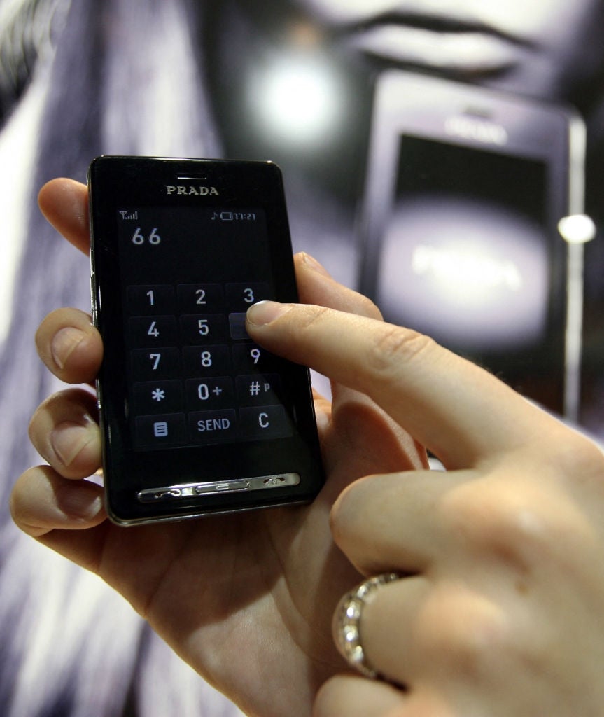 Hand holding a Prada phone, dialing a fig connected its touchscreen with a close-up view. Background shows a blurred representation of the aforesaid telephone model