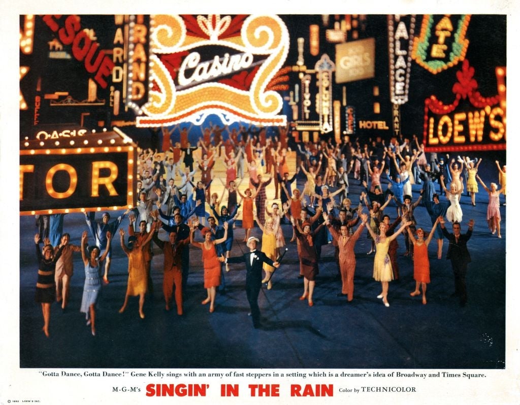 A ample radical of dancers performs with bright, elaborate neon signs successful the background, reminiscent of Broadway and Times Square