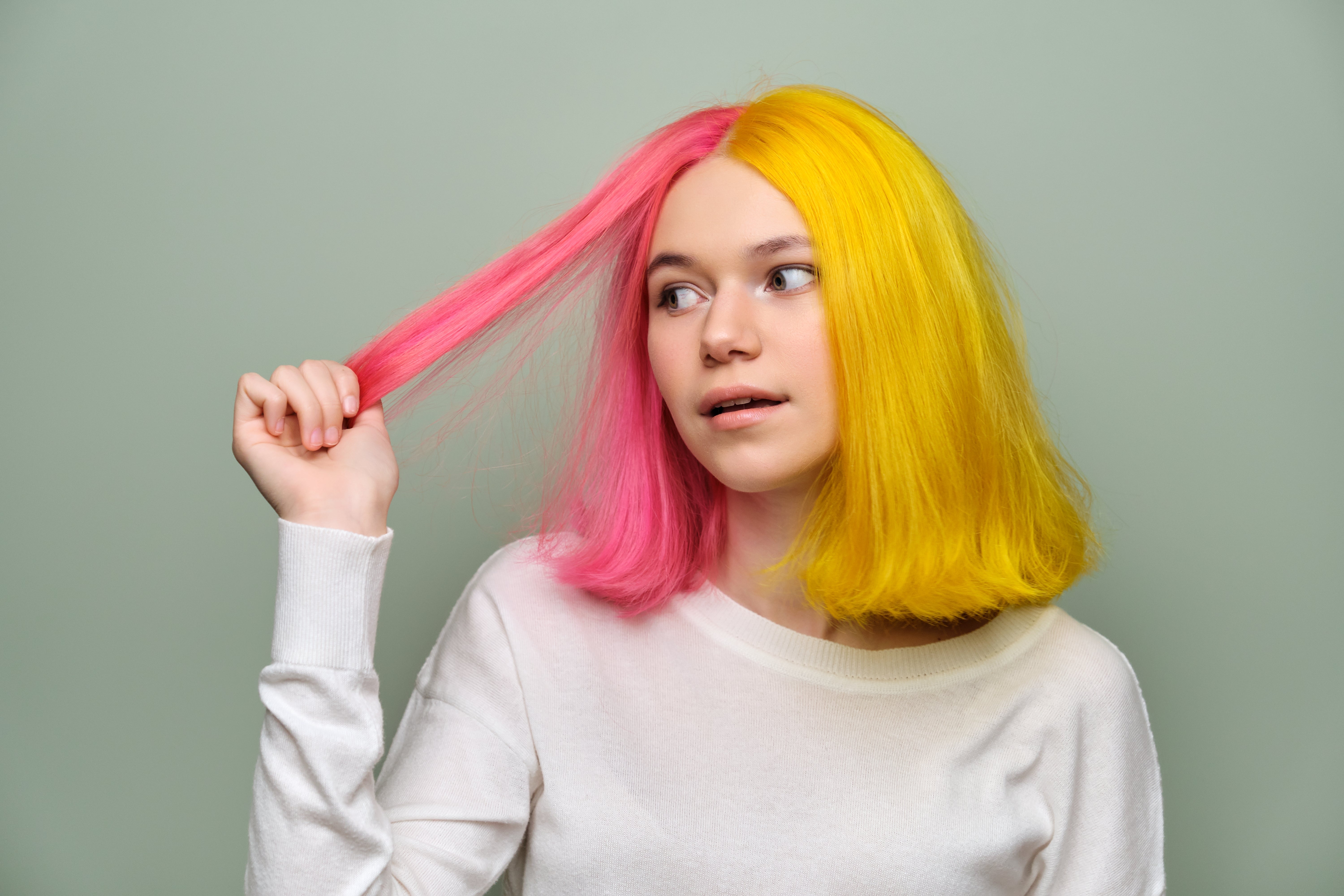 Person with split pink and yellow hair holding a strand, wearing a casual white shirt, looking to the side with a neutral expression