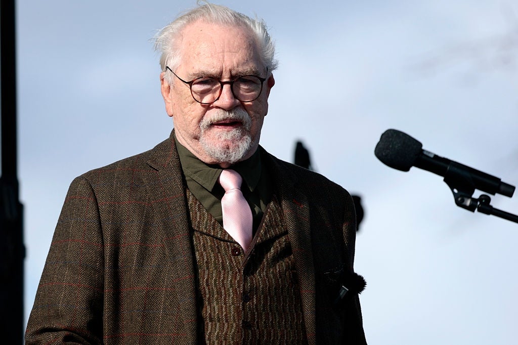 Eldre mann med briller og hvitt hår i brun rutete dress, mørk skjorte og rosa slips snakker på et utendørsarrangement med en mikrofon