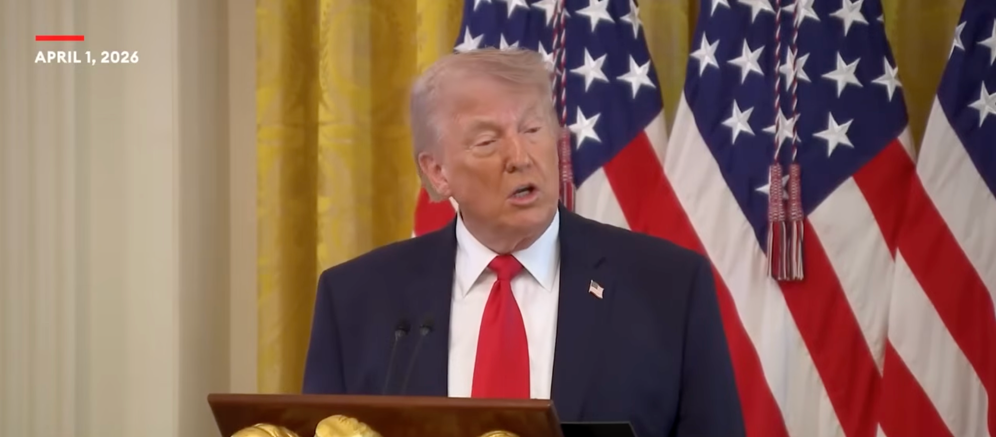 En person i dress snakker ved et podium med amerikanske flagg i bakgrunnen, datert 1. april 2026