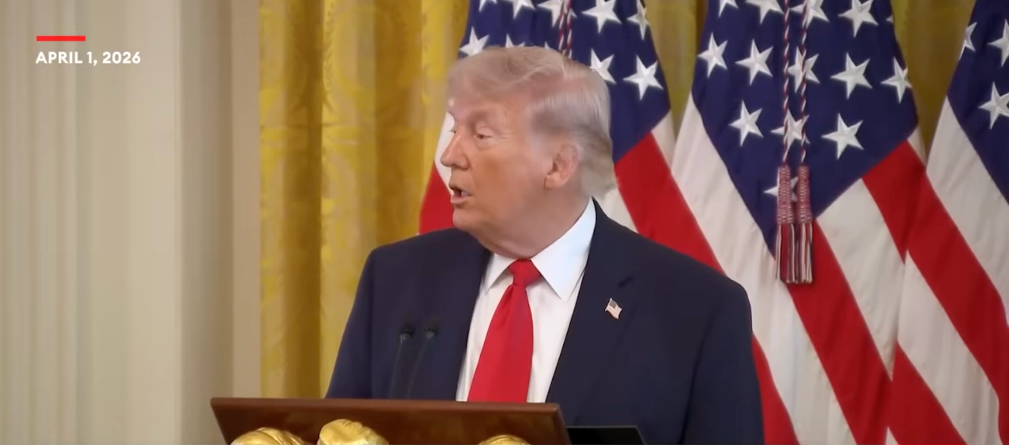 Mann snakker på et podium med USA-flagg i bakgrunnen. Dato: 1. april 2026, vises øverst i venstre hjørne