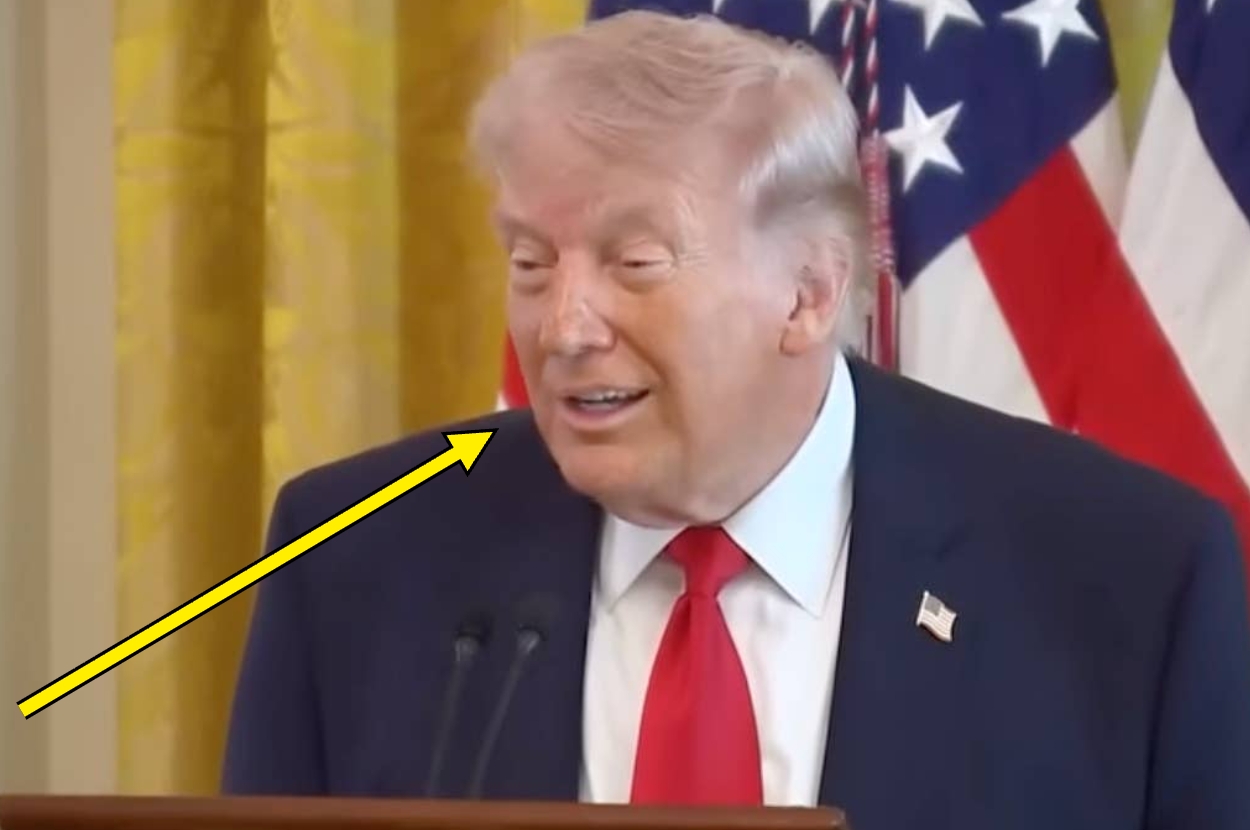 A person speaking at an event, wearing a suit and tie, with flags in the background