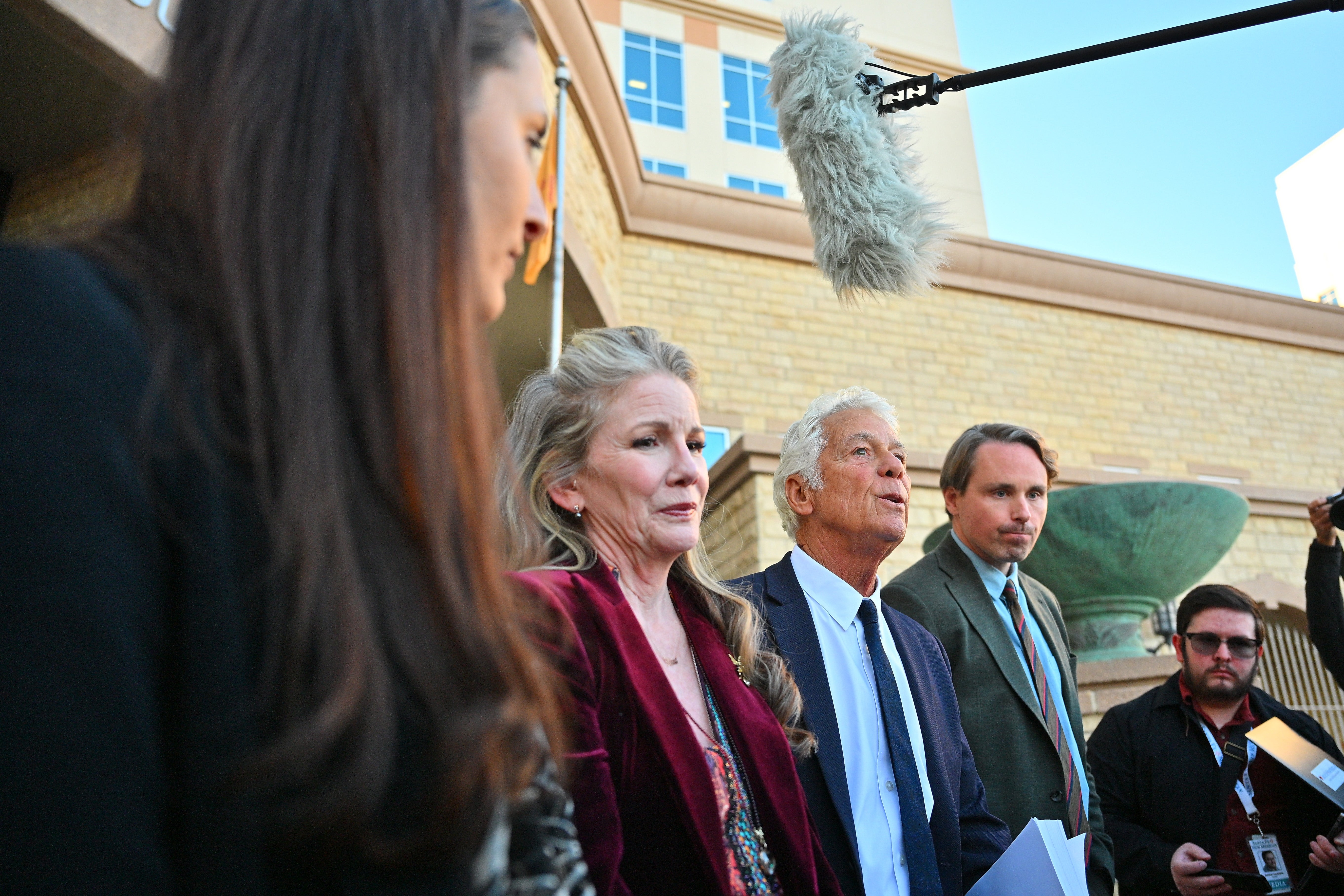 Gruppe mennesker, inkludert en eldre mann i dress og en eldre kvinne i fløyelsantrekk, snakker til pressen utenfor en bygning med en mikrofon over hodet