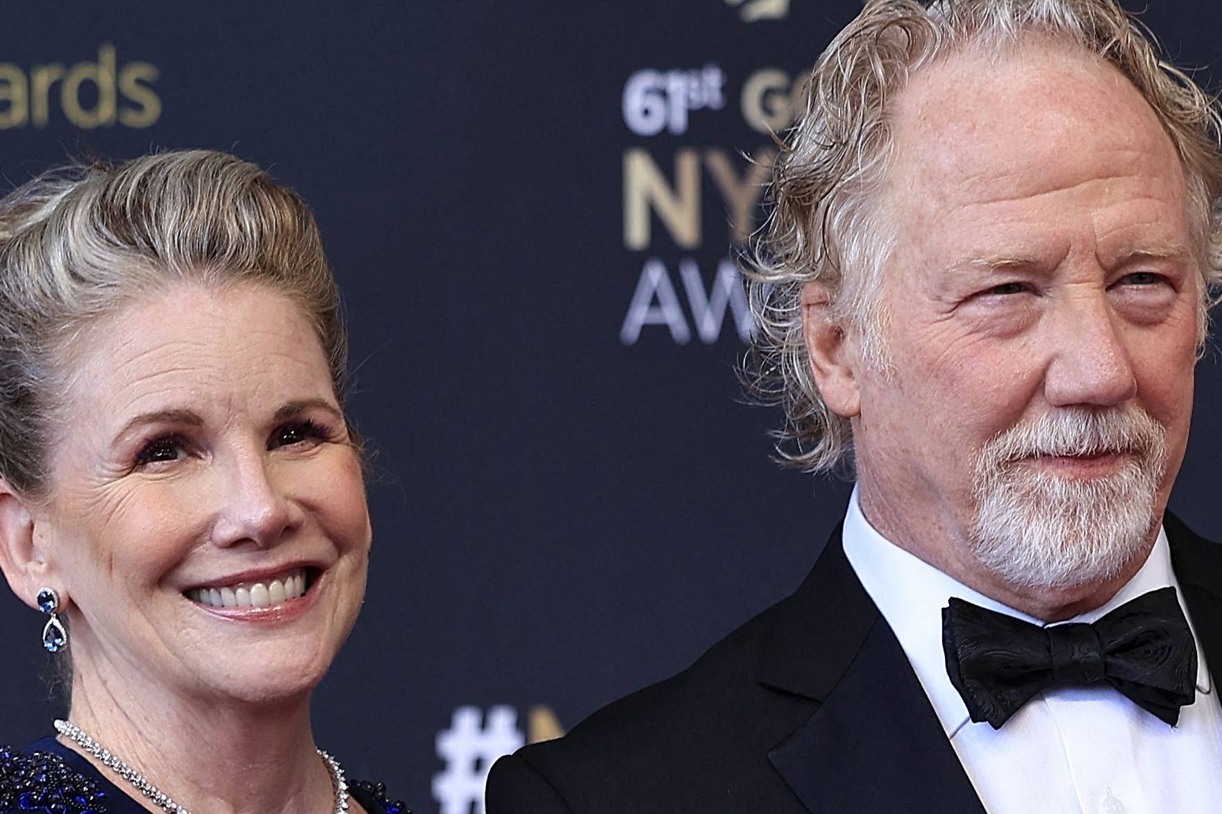Two people on a red carpet. One wears a formal gown with cape-like sleeves, the other a classic tuxedo. They smile while posing for photos
