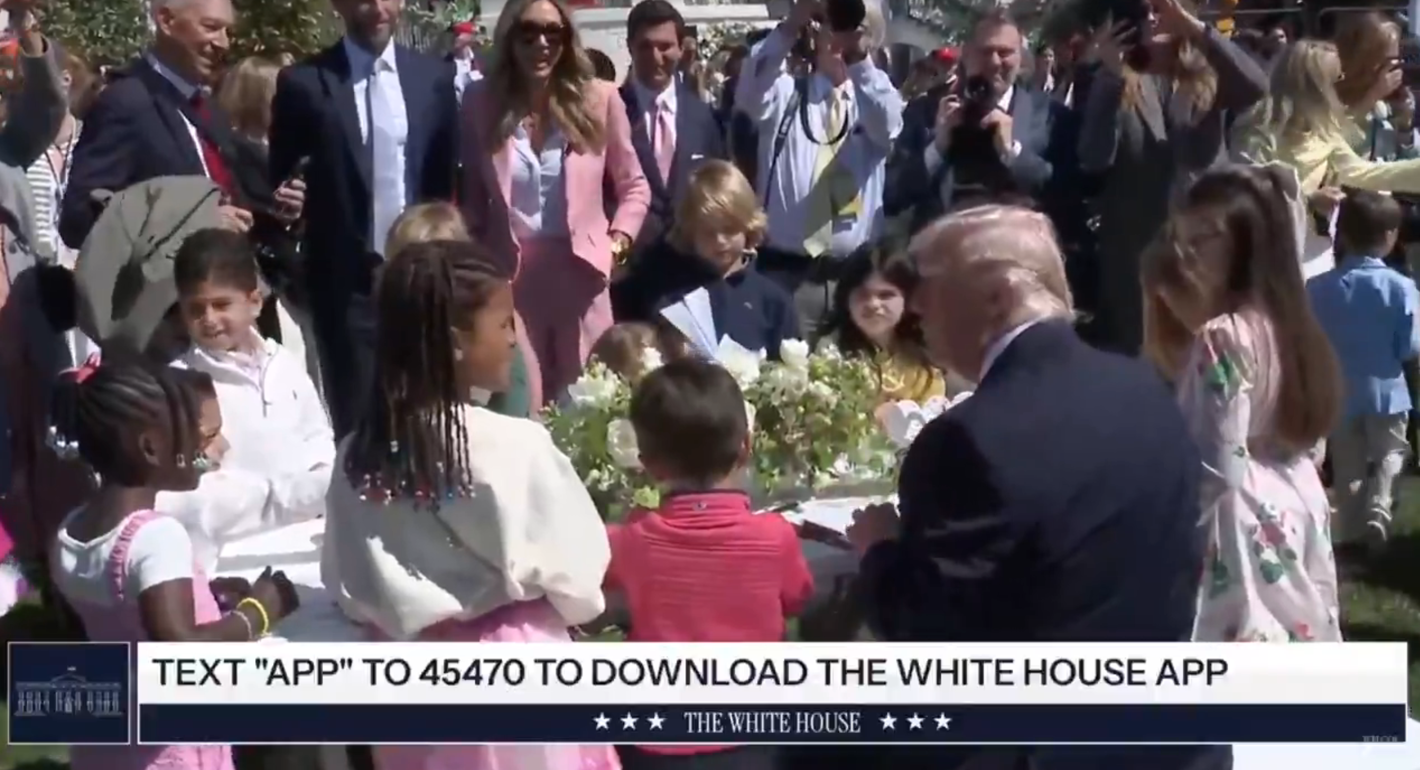 Barn samles rundt et bord med blomster mens voksne ser på en offentlig begivenhet. En promo for Det hvite hus-appen vises under scenen