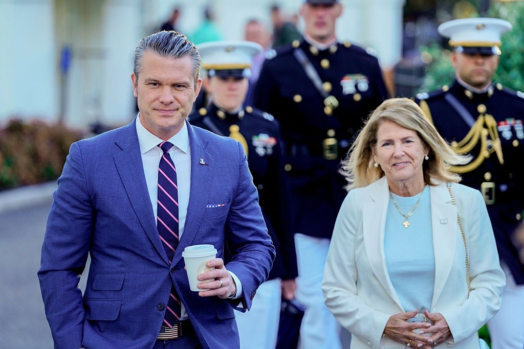 En mann i dress og slips holder en kaffekopp. En kvinne ved siden av ham har på seg en hvit dress. De går foran uniformert personell