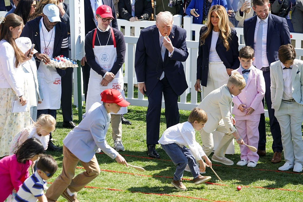 En gruppe barn deltar i et utendørs eggrace-arrangement som voksne, inkludert enkeltpersoner i formell antrekk, ser og heier på dem