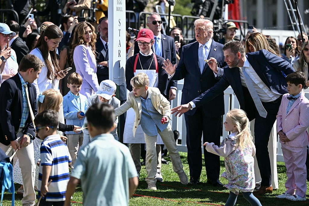 Folk, inkludert barn, er samlet utendørs for et påskeeggrullarrangement, med en mann i dress som oppmuntrer et barn til aktiviteten