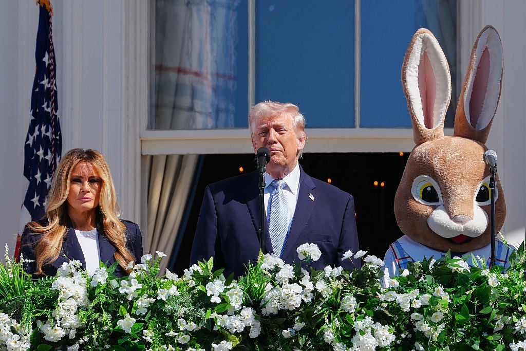 Tre personer står på en balkong: en kvinne i et elegant antrekk, en mann i dress som snakker ved et podium, og en påskehare-maskot