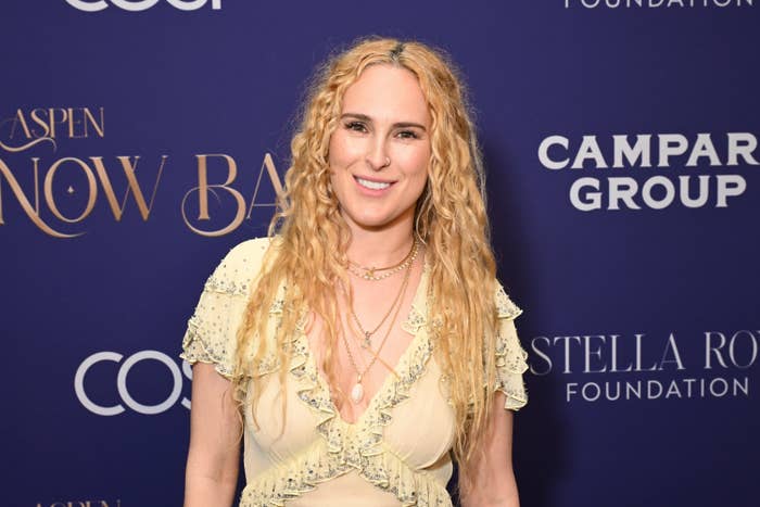 Person connected  reddish  carpet successful  elegant, ruffled gown with layered necklaces, smiling against backdrop with lawsuit   logos