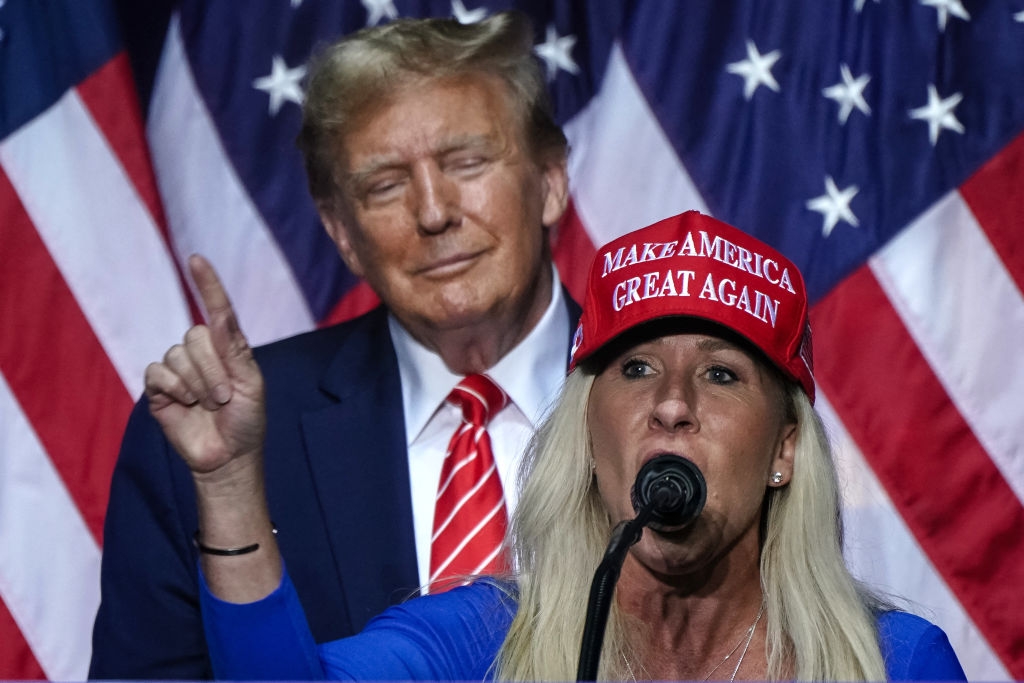 A idiosyncratic   successful  a "Make America Great Again" chapeau  speaks astatine  a podium, with an American emblem  backdrop and different  idiosyncratic  successful  the background