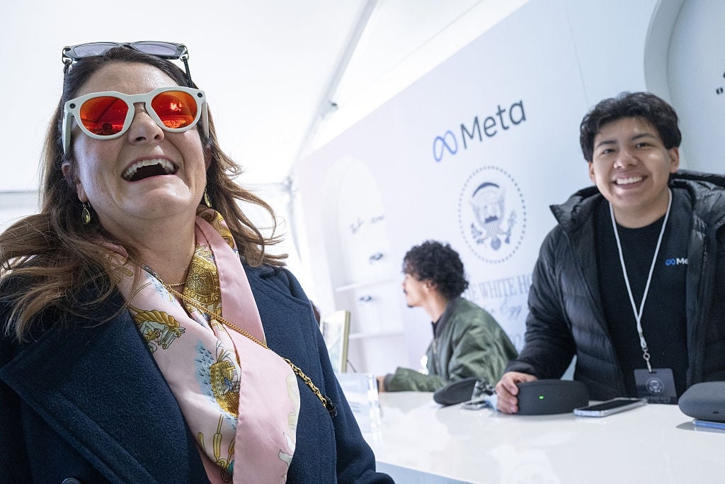 Kvinne i stilige solbriller og skjerf smiler bredt på en Meta-eventstand; to menn samhandler i bakgrunnen