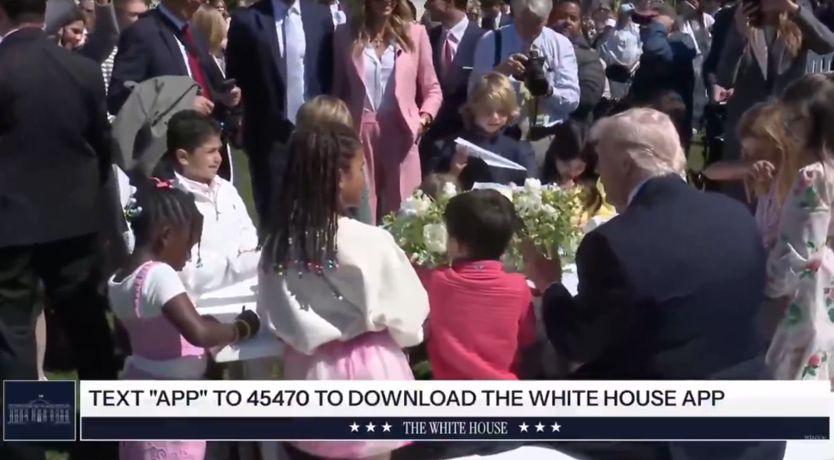 Folkemengden samles rundt et utendørs bord; barnesetesenter med andre stående. Tekstmelding: "Tekst 'APP' til 45470 for å laste ned Det hvite hus-appen."