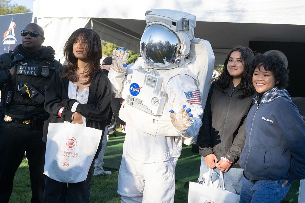En gruppe poserer med en person i en astronautdress utendørs, sammen med en sikkerhetsoffiser. Man holder en minnesekk