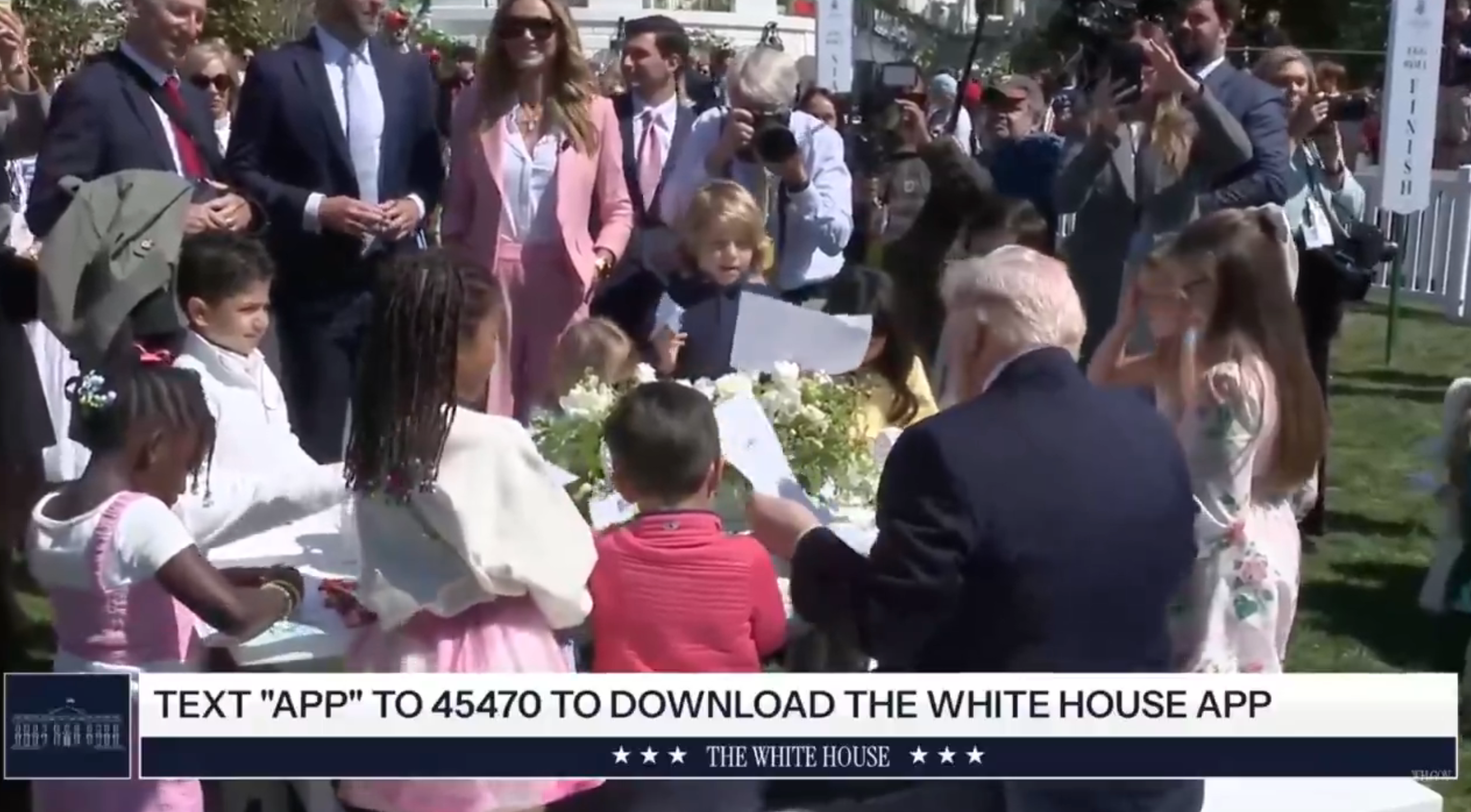 Barn samlet rundt et bord med en offisiell som delte ut gjenstander. Tekst nederst fremmer nedlasting av Det hvite hus-appen