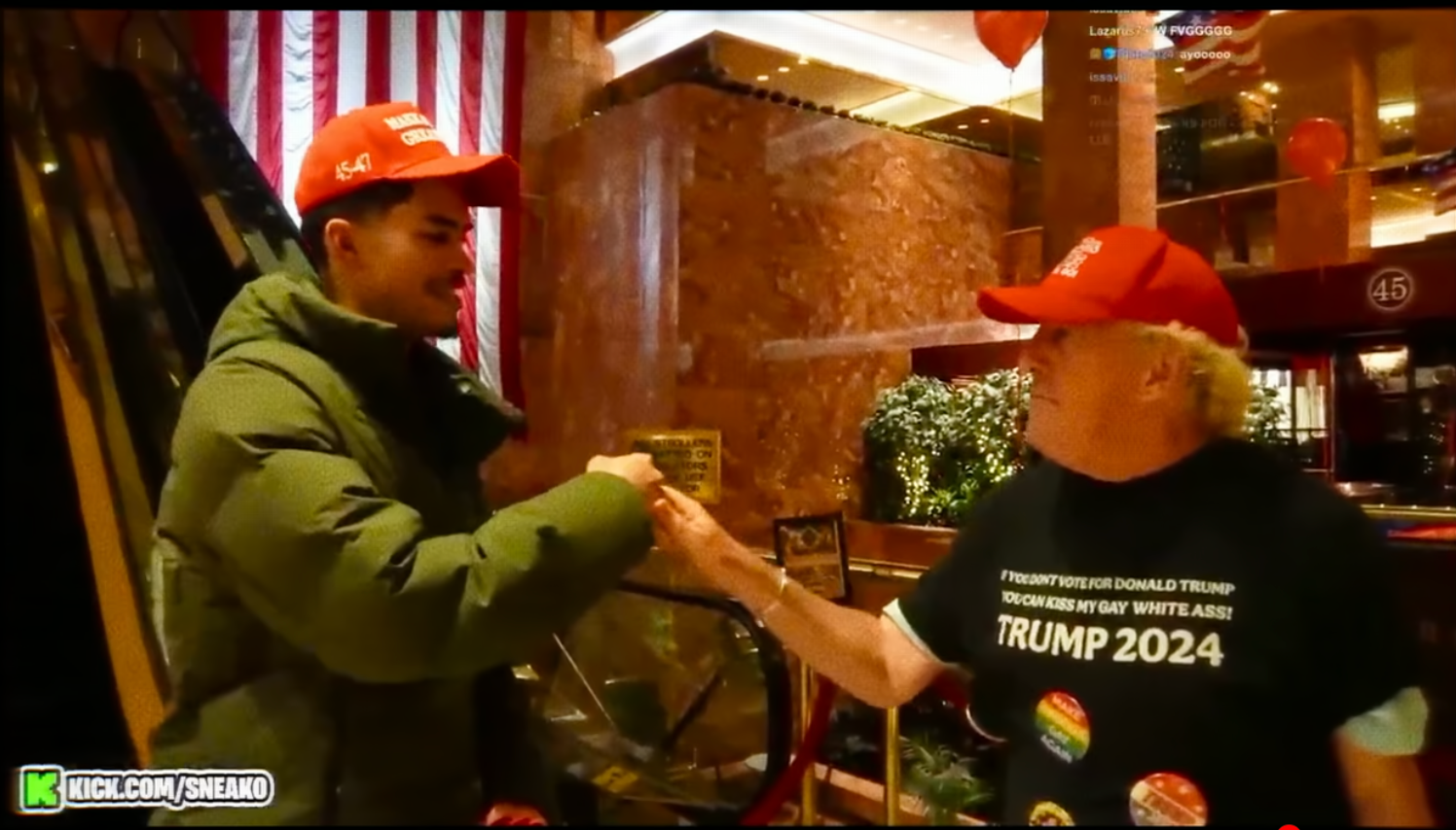 Two radical wearing reddish caps speech a fist bump indoors. One idiosyncratic wears a greenish puffer jacket, and the different a achromatic T-shirt with governmental text